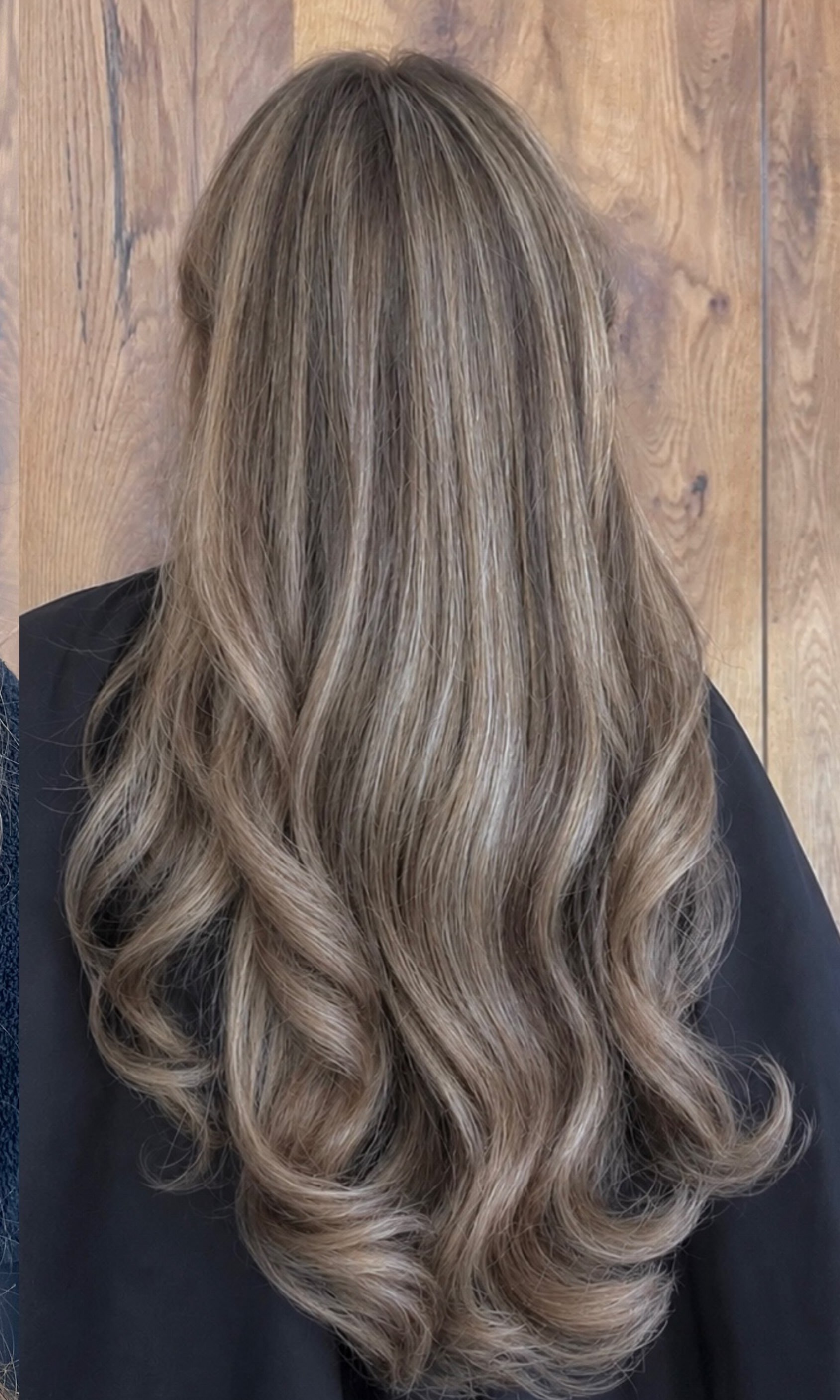 Lang haar met een asblonde balayage in zachte golven, van achteren gezien voor de houten wand van de salon